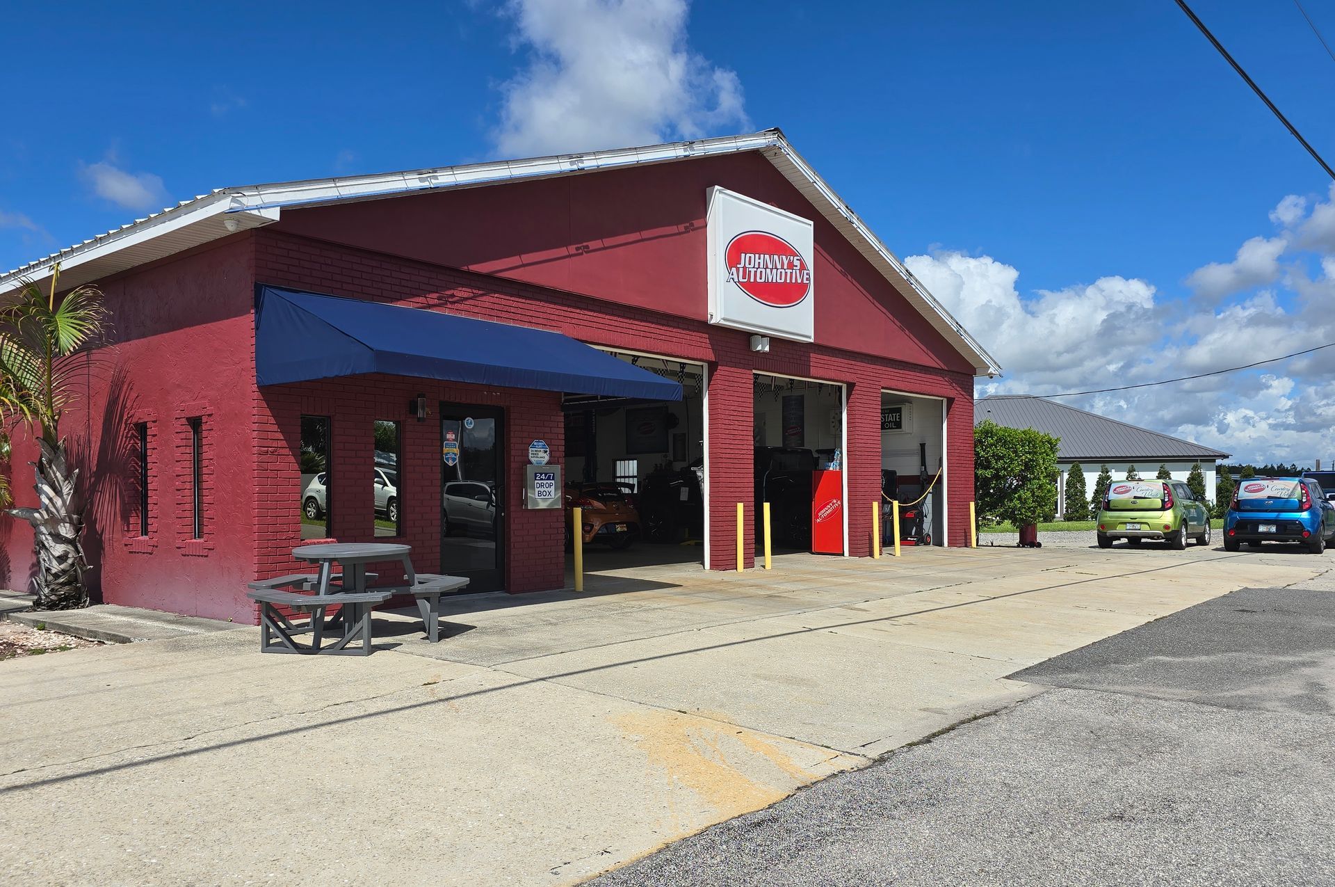 Red auto repair shop with blue awning and sign, vehicles parked outside on sunny day. | Johnny's Automotive