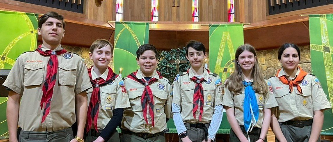 A group of six people in Boy Scout uniforms stand in front of a wood background.