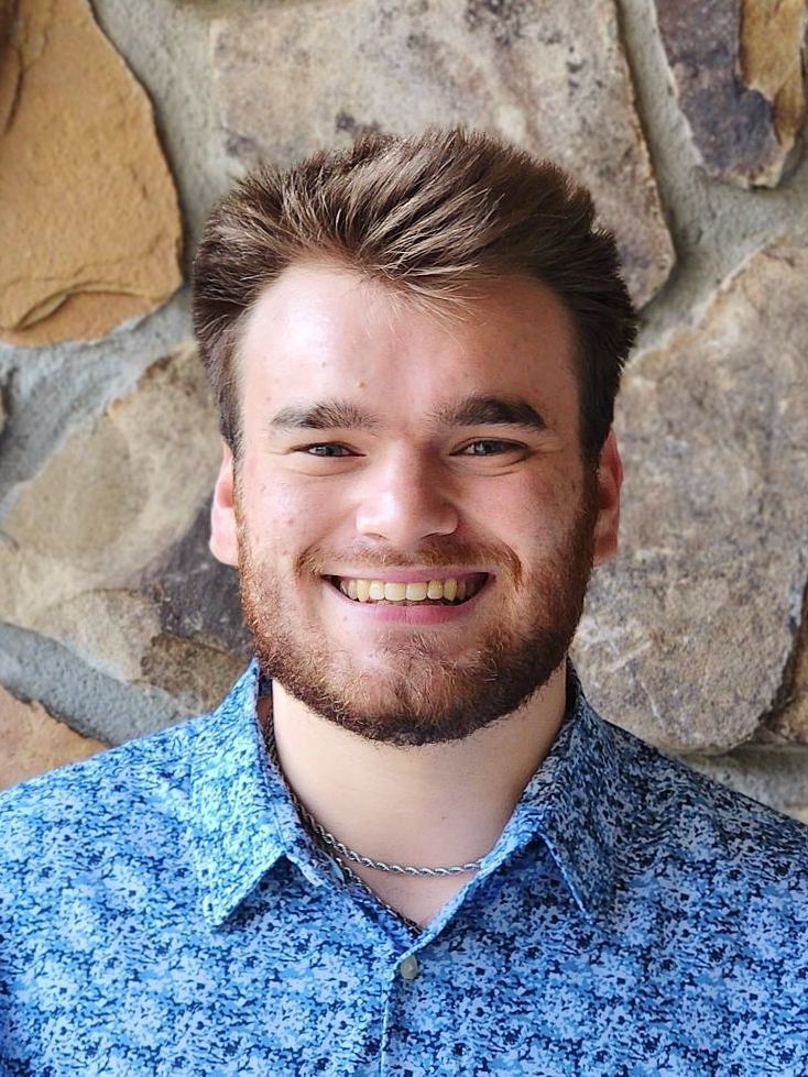 Man with brown hair and beard smiles, wearing a blue patterned shirt. Stone wall background.