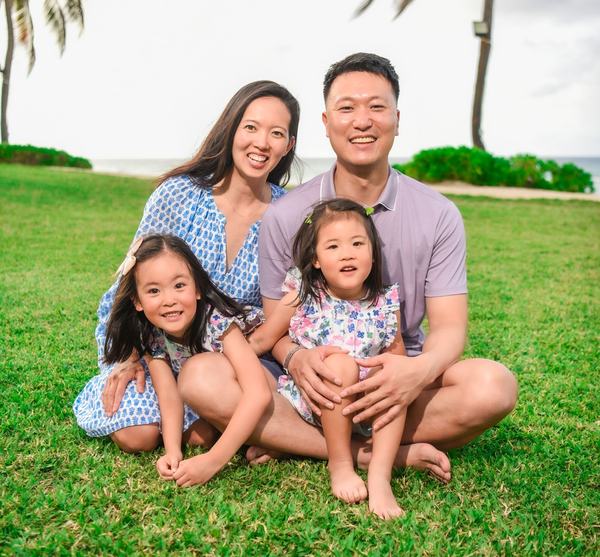 Family of four seated on green grass; woman in blue dress, man in purple shirt, two young girls.