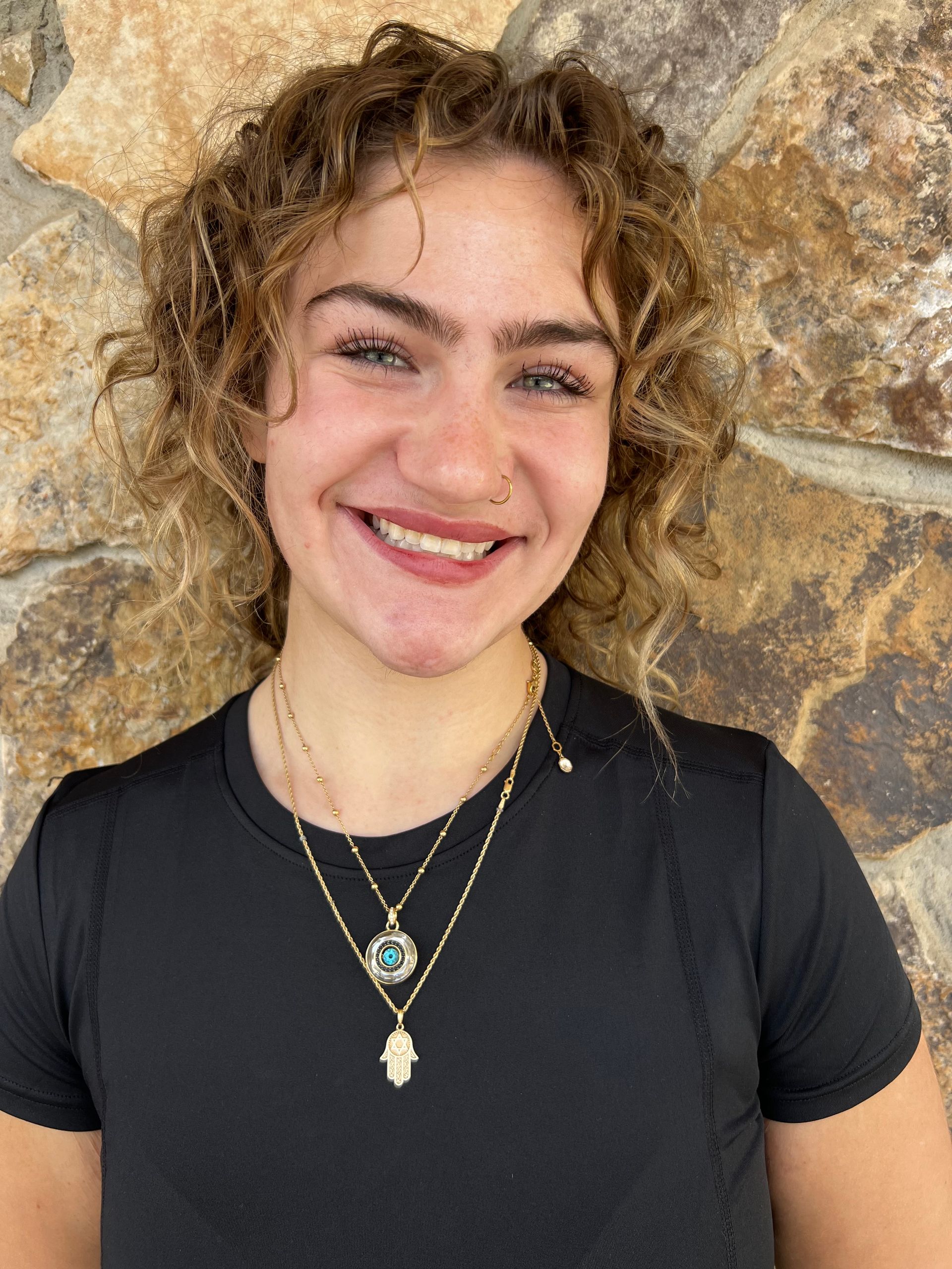 Woman with blonde hair smiling, wearing a white t-shirt, standing in front of a stone wall.