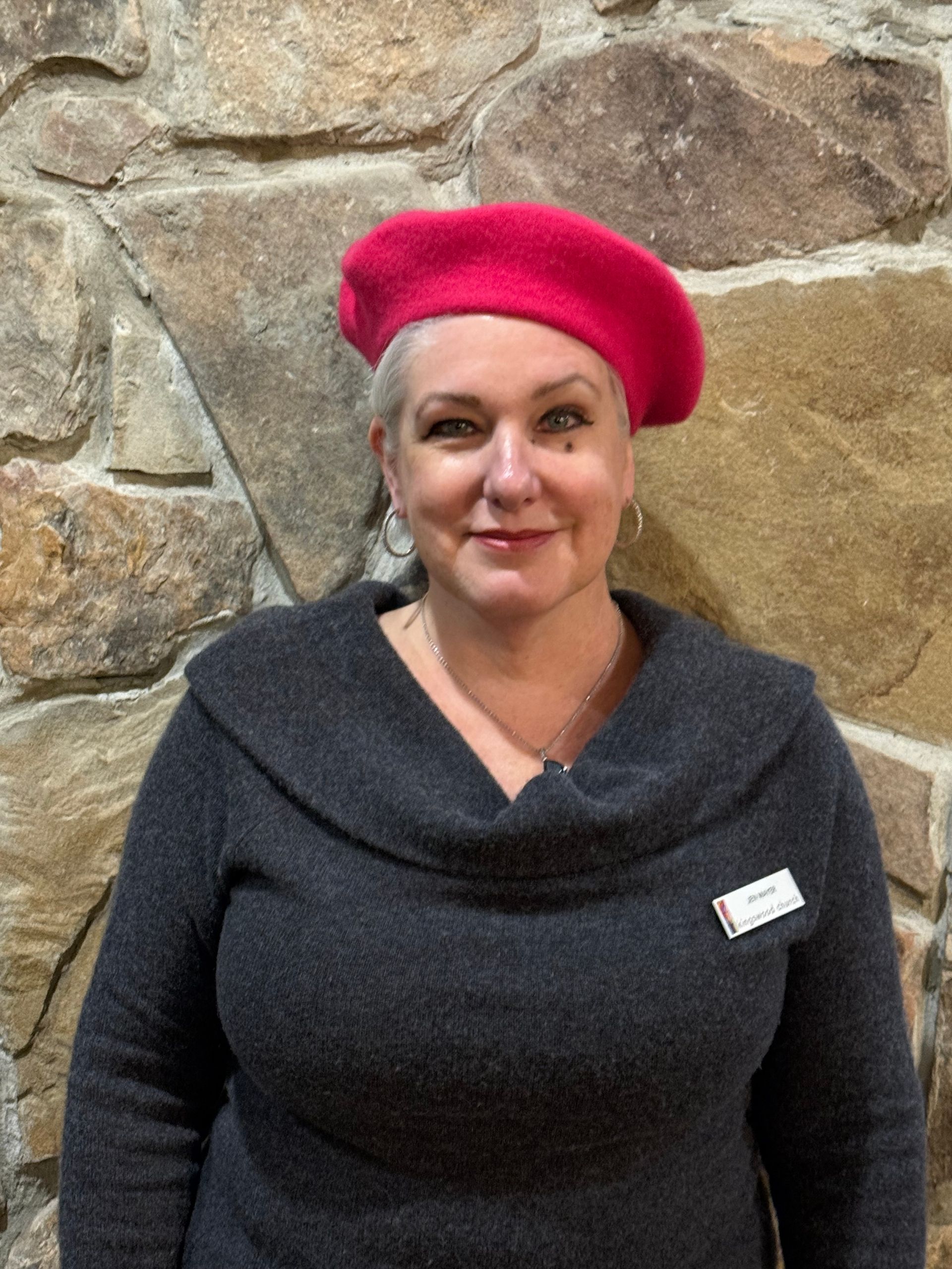 Woman wearing a red beret and gray sweater smiles in front of a stone wall.