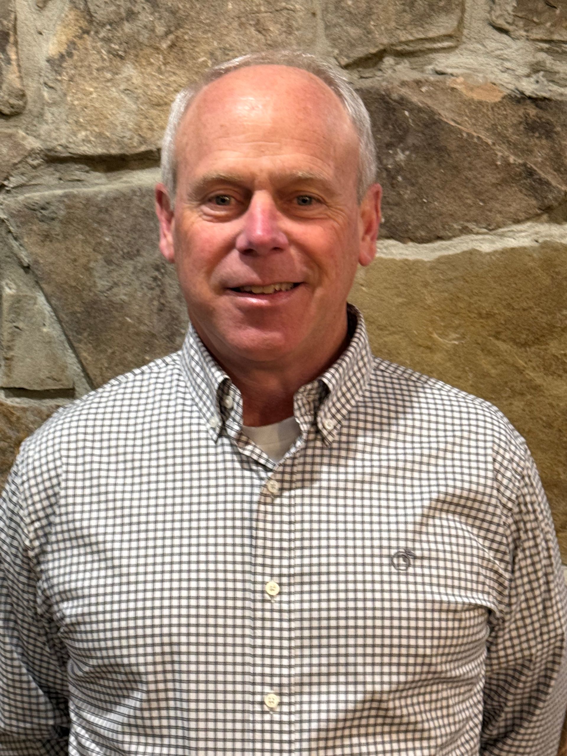 Man in a plaid button-down shirt smiles, standing in front of a stone wall.