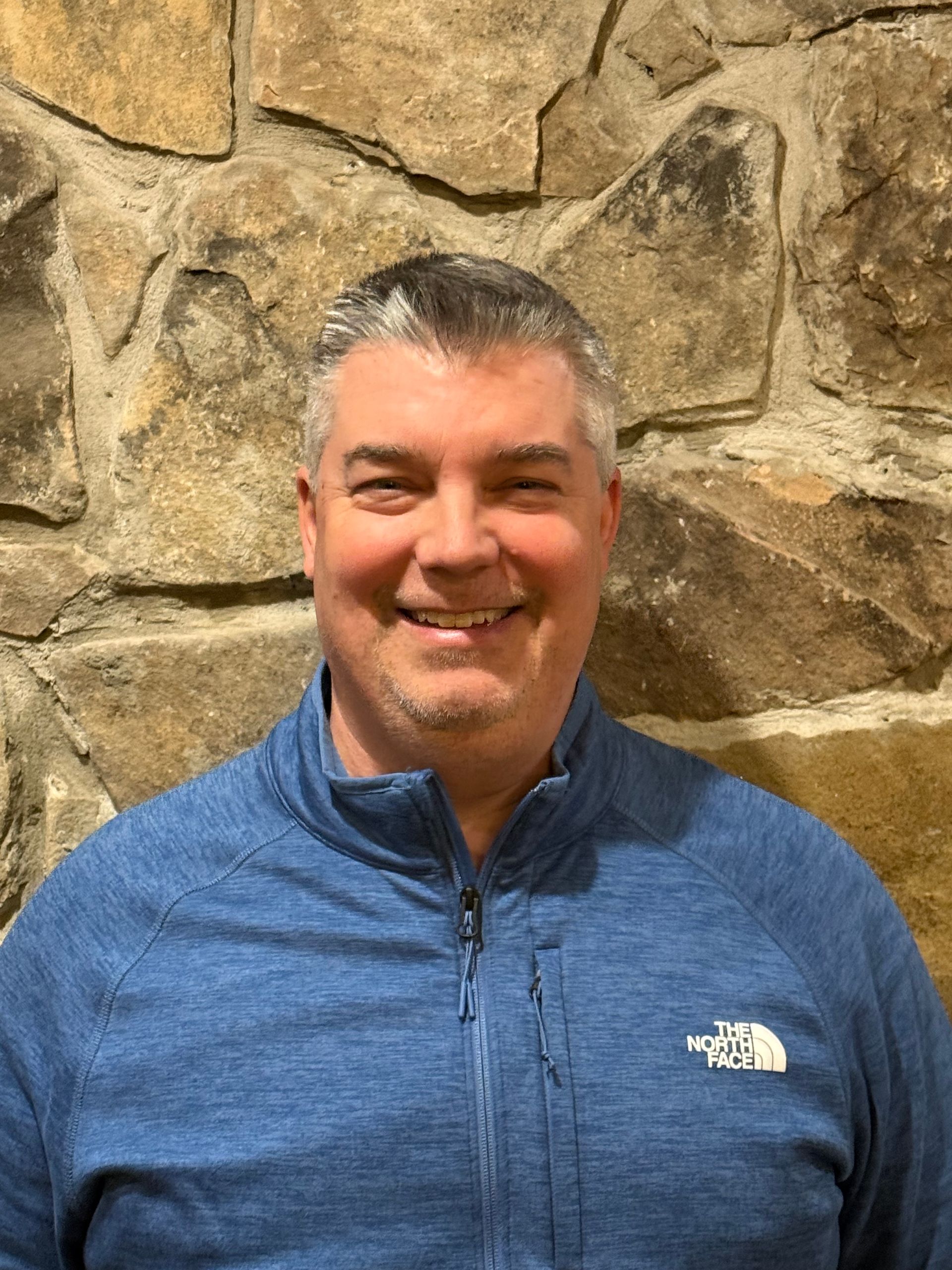 Man in blue zip-up smiling in front of a stone wall.