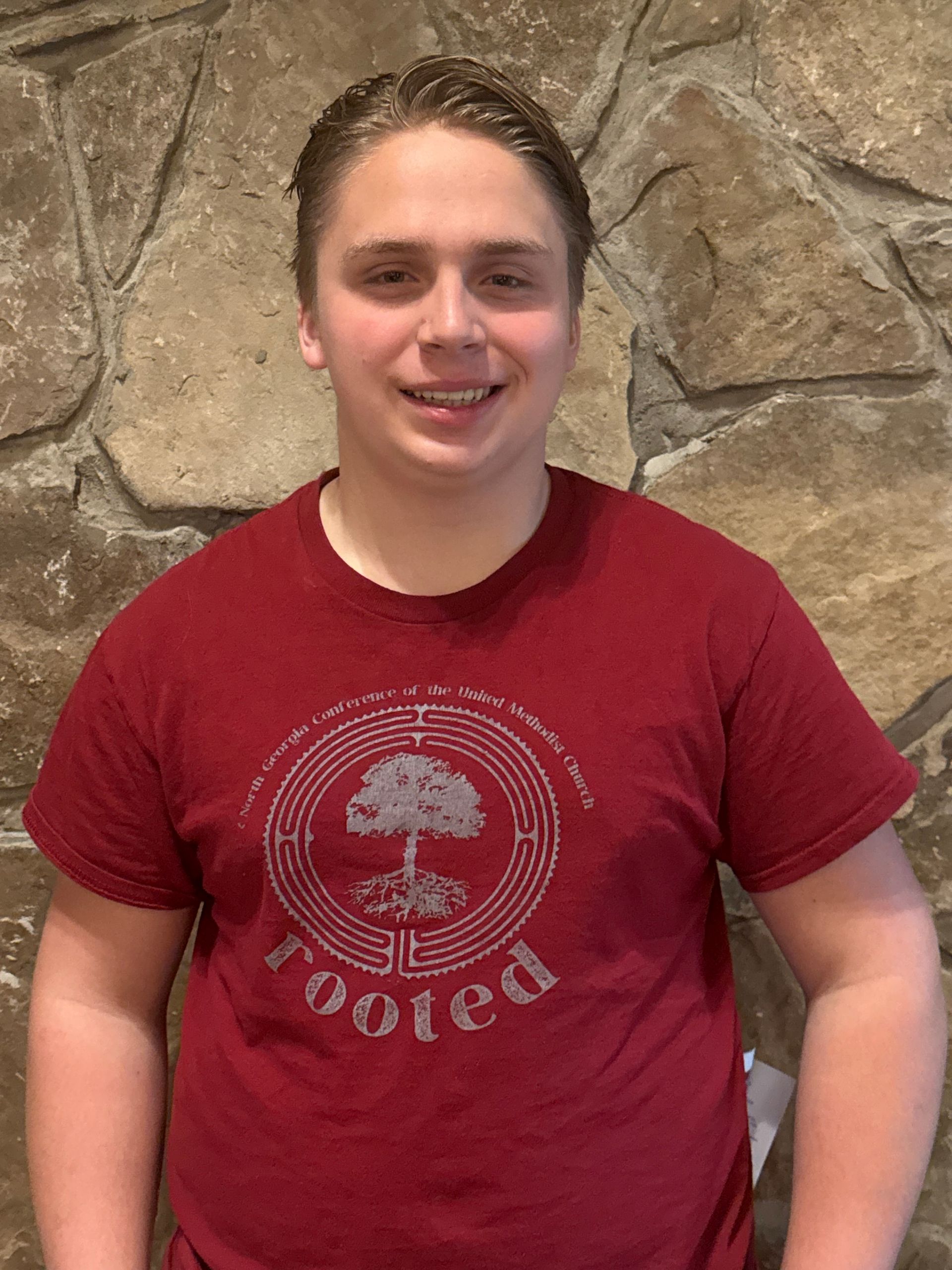 Woman with dark hair smiles, wearing a Kingswood Church t-shirt, standing in front of a stone wall.