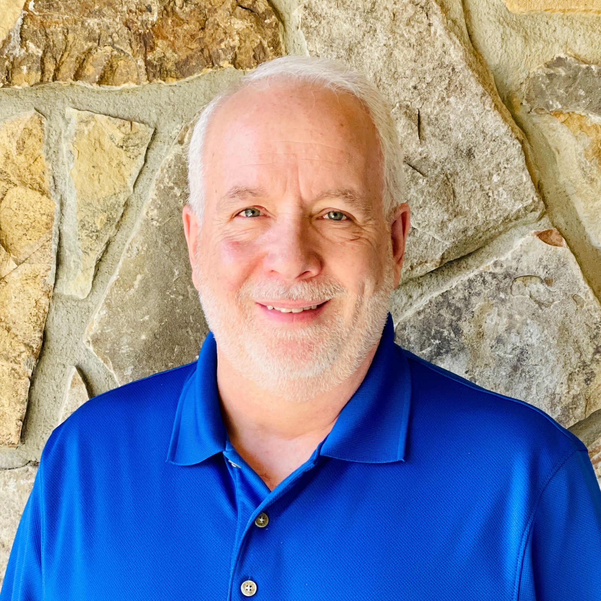 Man with short white hair and beard smiles, wearing a blue polo shirt, standing in front of a stone wall.