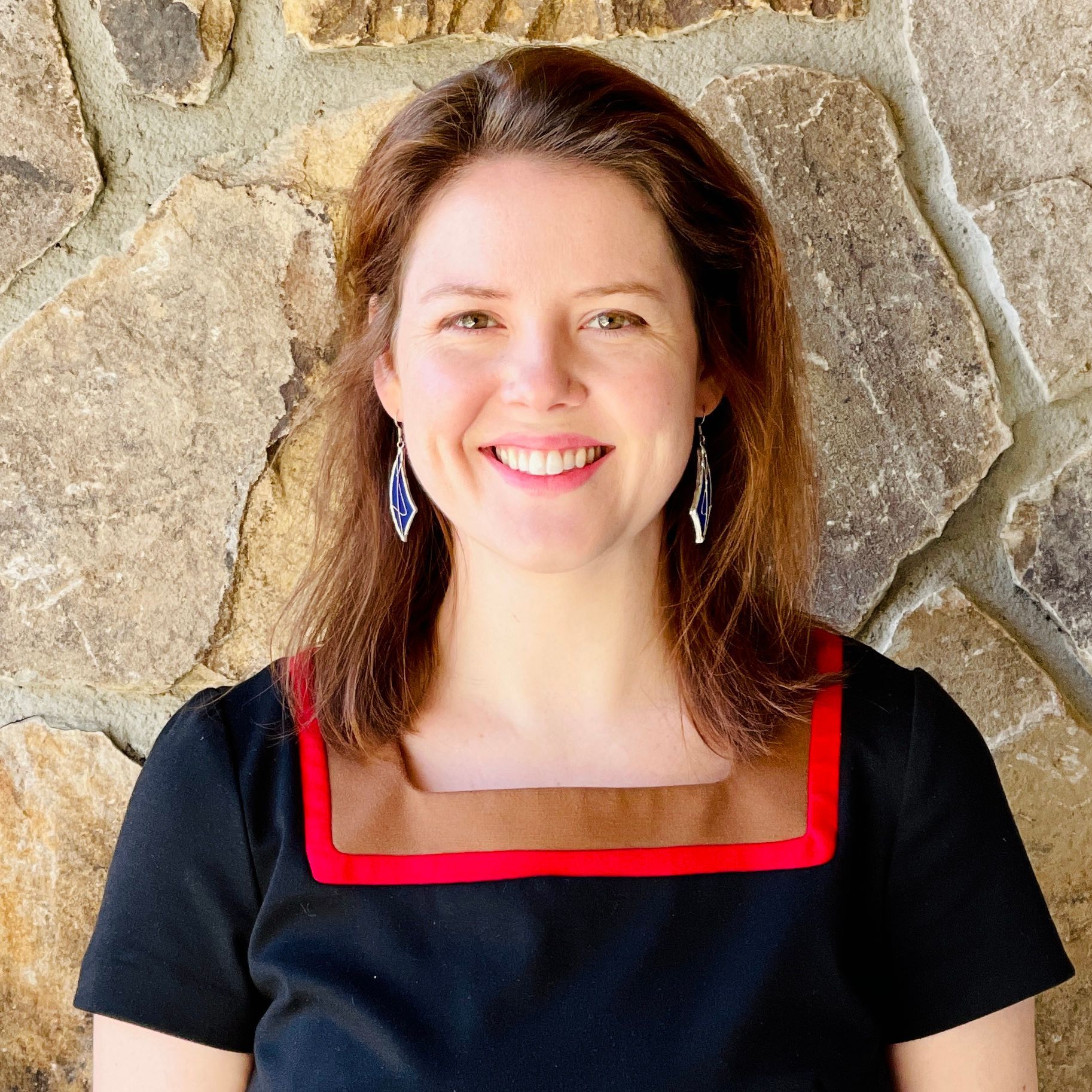 Woman with brown hair smiles, wearing a black dress with a brown and red square neckline, in front of a stone wall.