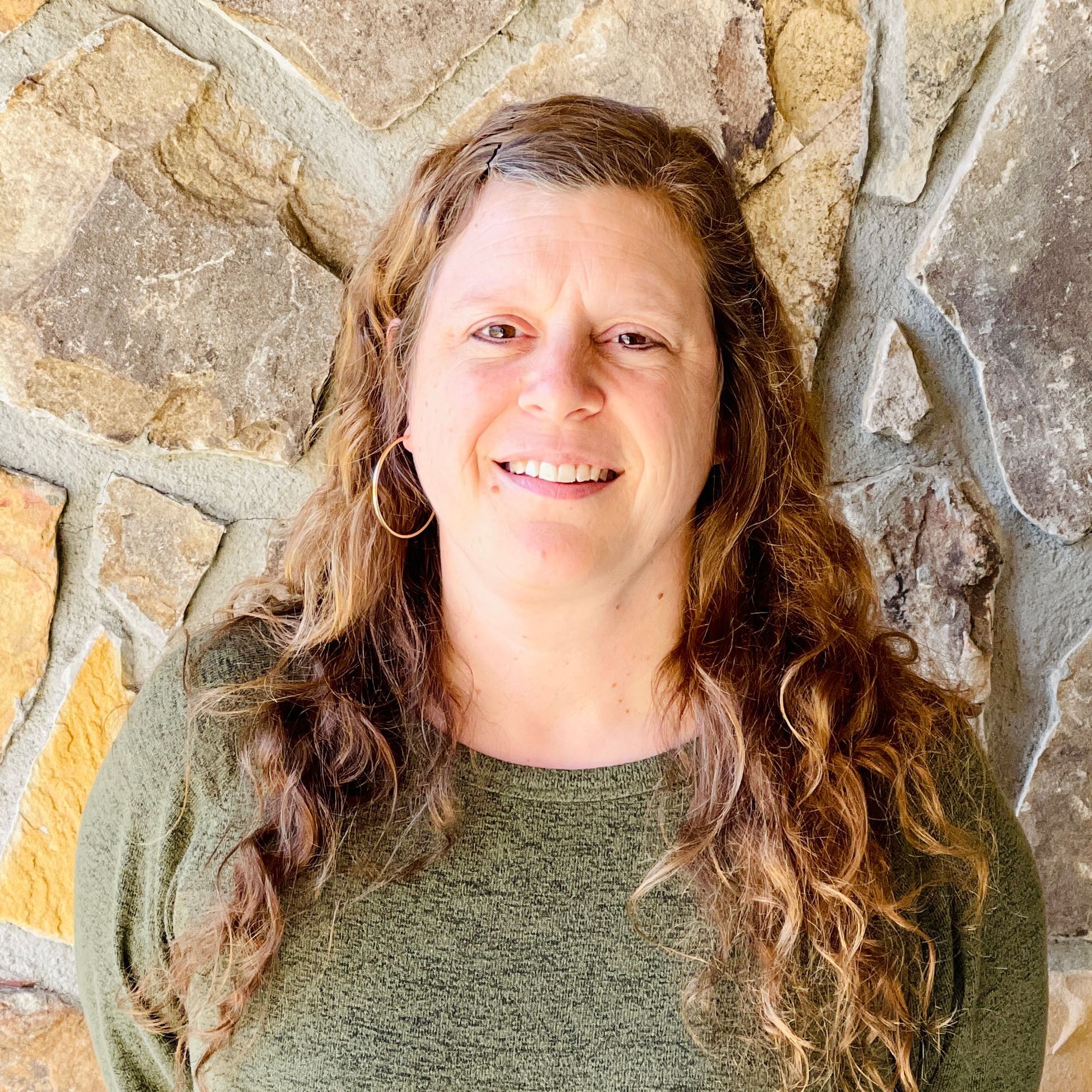 Woman with long, wavy brown hair smiles in front of a stone wall. She wears a green sweater and hoop earrings.