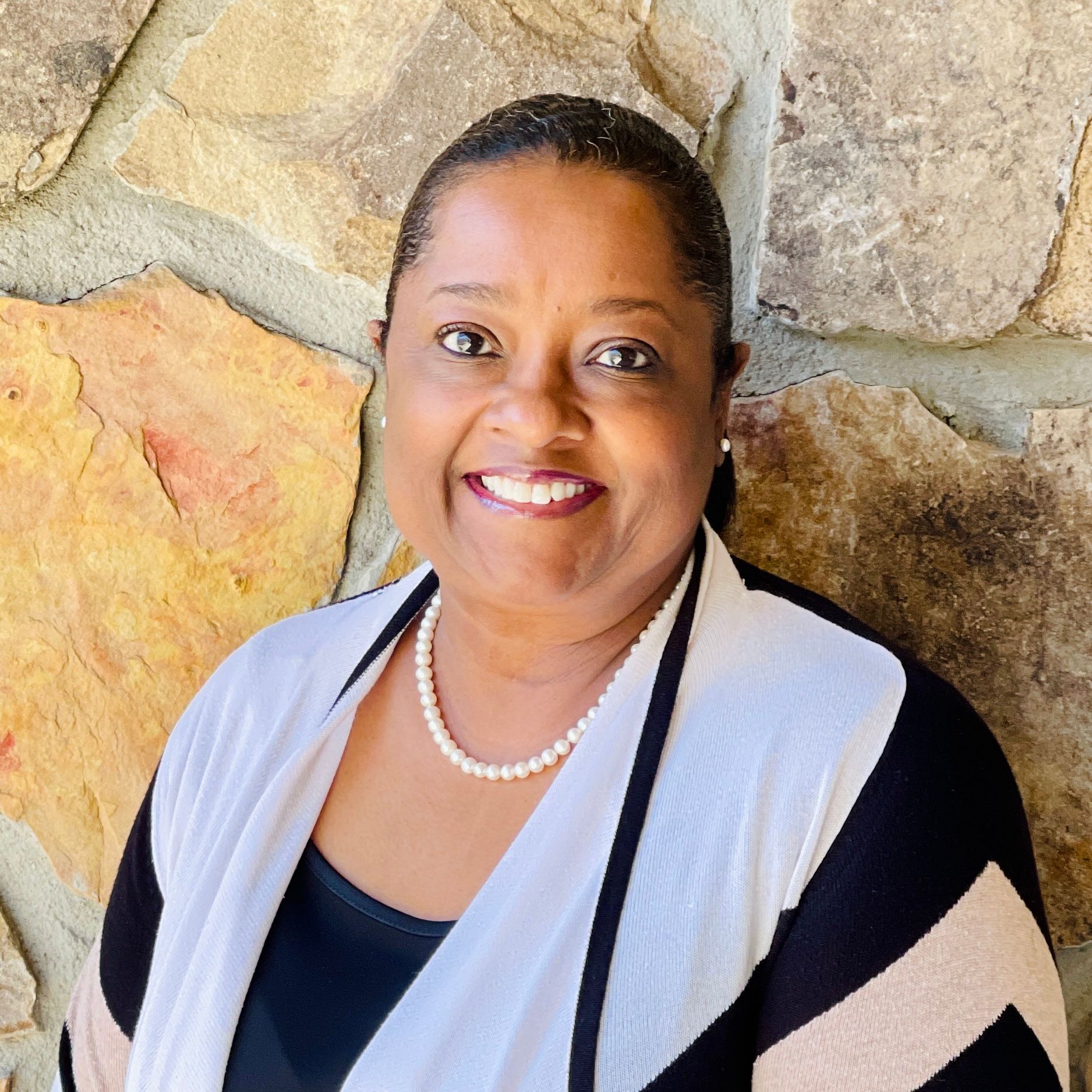 Woman smiling, wearing a pearl necklace, black top and cardigan, standing in front of a stone wall.