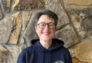 Woman in glasses and navy blue hooded sweatshirt smiling in front of a stone wall.