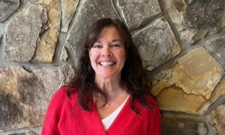 Woman with brown hair smiles, wearing a red sweater and white shirt, stands in front of a stone wall.
