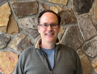 Man with glasses smiles, standing in front of a stone wall, wearing a zip-up jacket.