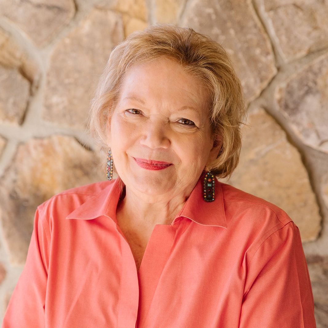 Woman with short, curly blonde hair smiles, wearing an orange collared shirt, stone background.