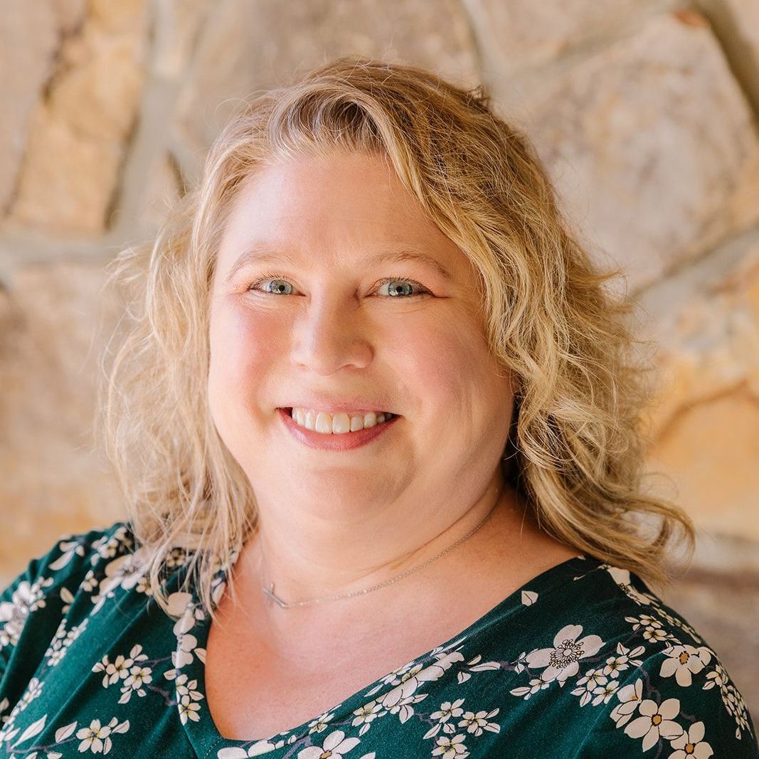 Woman with blonde curly hair smiles at the camera, wearing a green floral top, stone wall background.