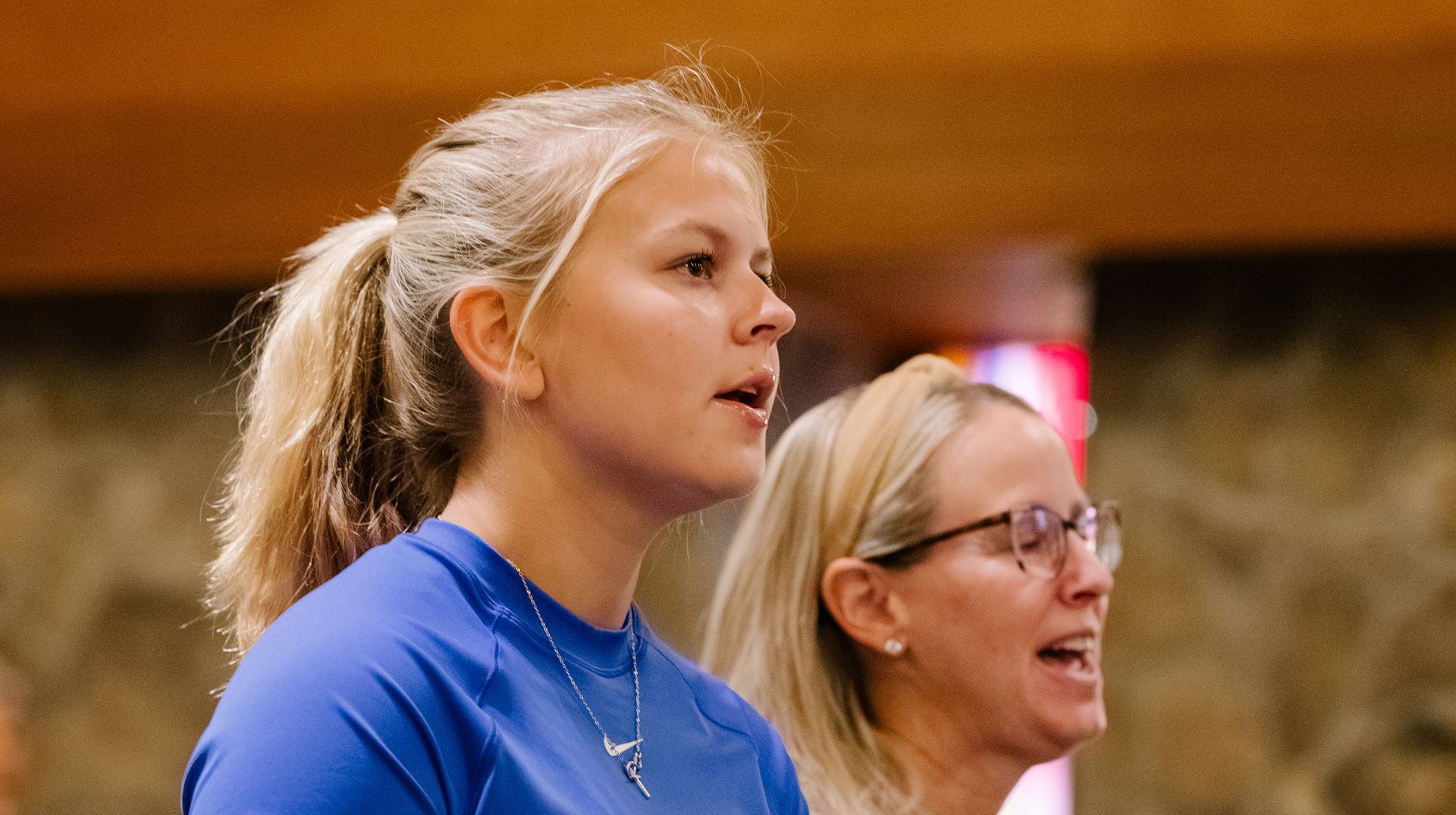 Two women singing in a church, one with blonde hair in a ponytail and a blue shirt, the other wearing glasses.