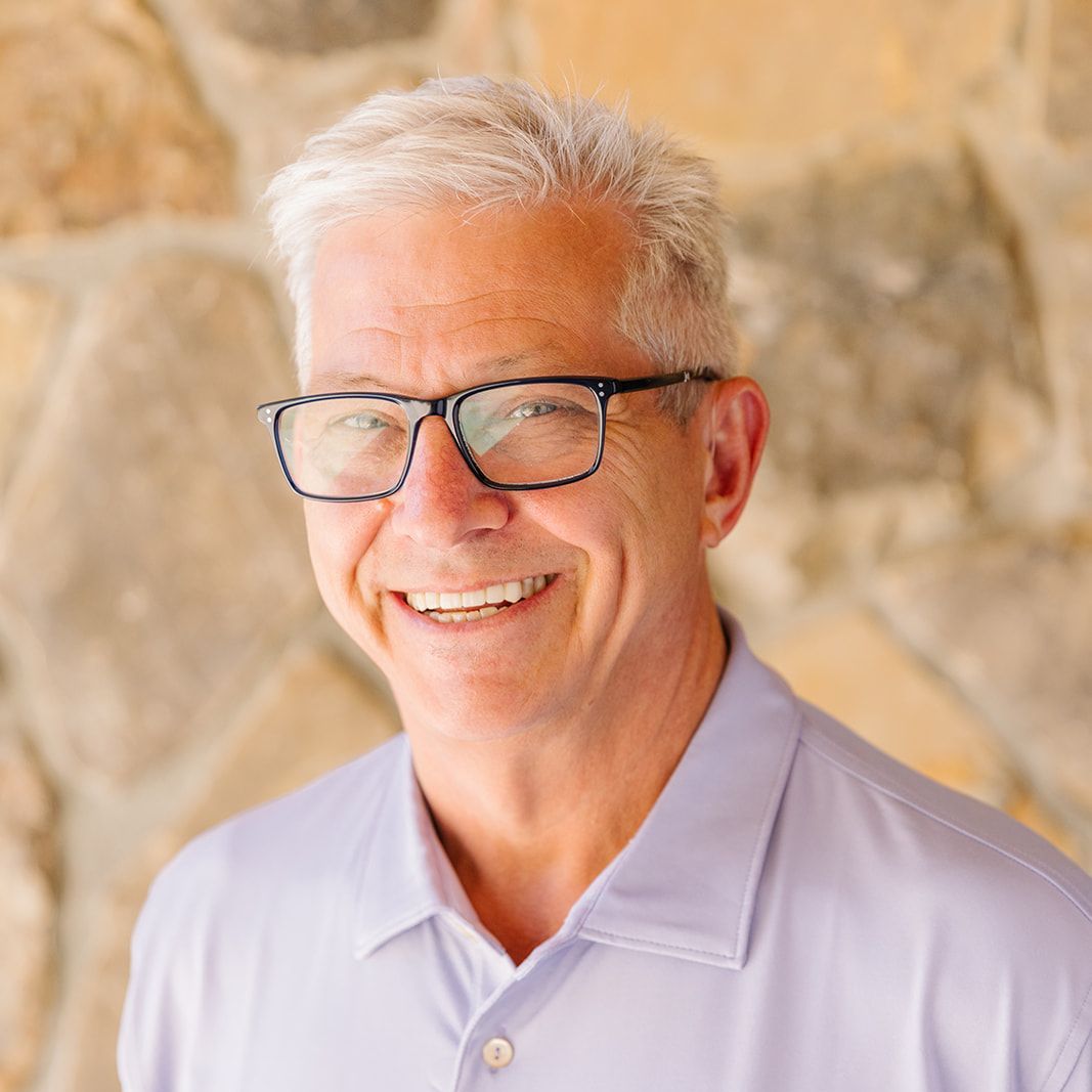 Man wearing glasses smiles, standing in front of a stone wall, wearing a purple polo shirt.