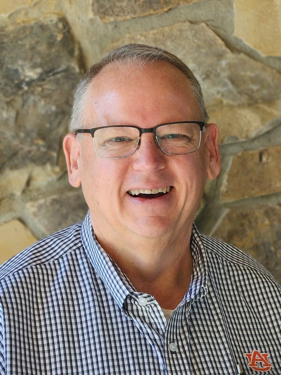 Man with glasses smiles, wearing yellow shirt, standing in front of stone wall.