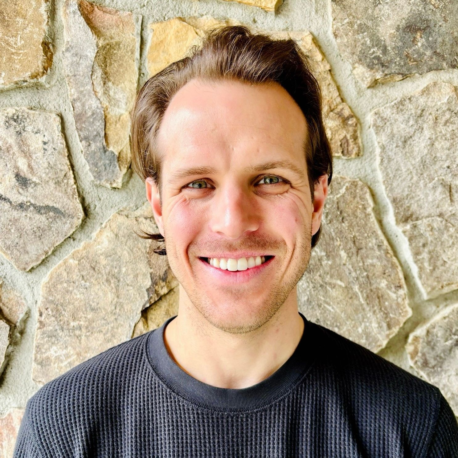 Man smiling, facing forward, against a stone wall background. Wearing a dark sweater.
