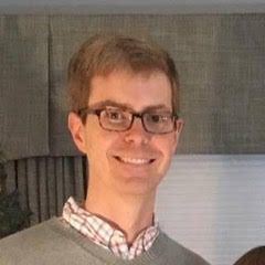 Man wearing glasses smiles, in a light gray sweater and plaid shirt.