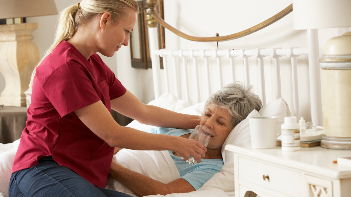 Nurse in red scrubs giving water to a sick elderly woman in bed.