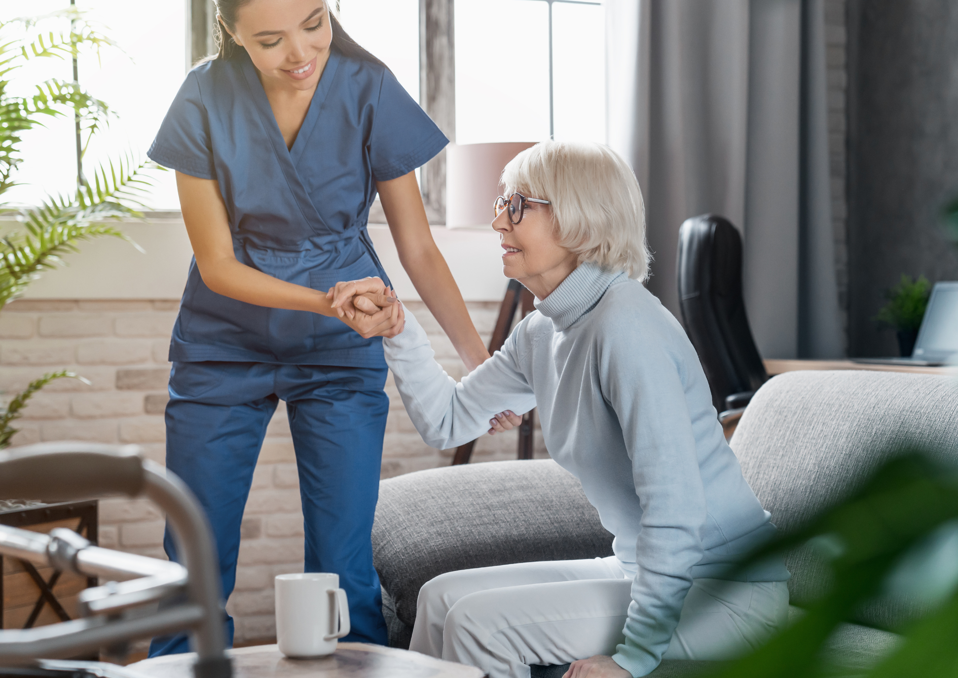 Caregiver in blue scrubs helps an older woman in a light blue sweater stand up from a sofa in a home.