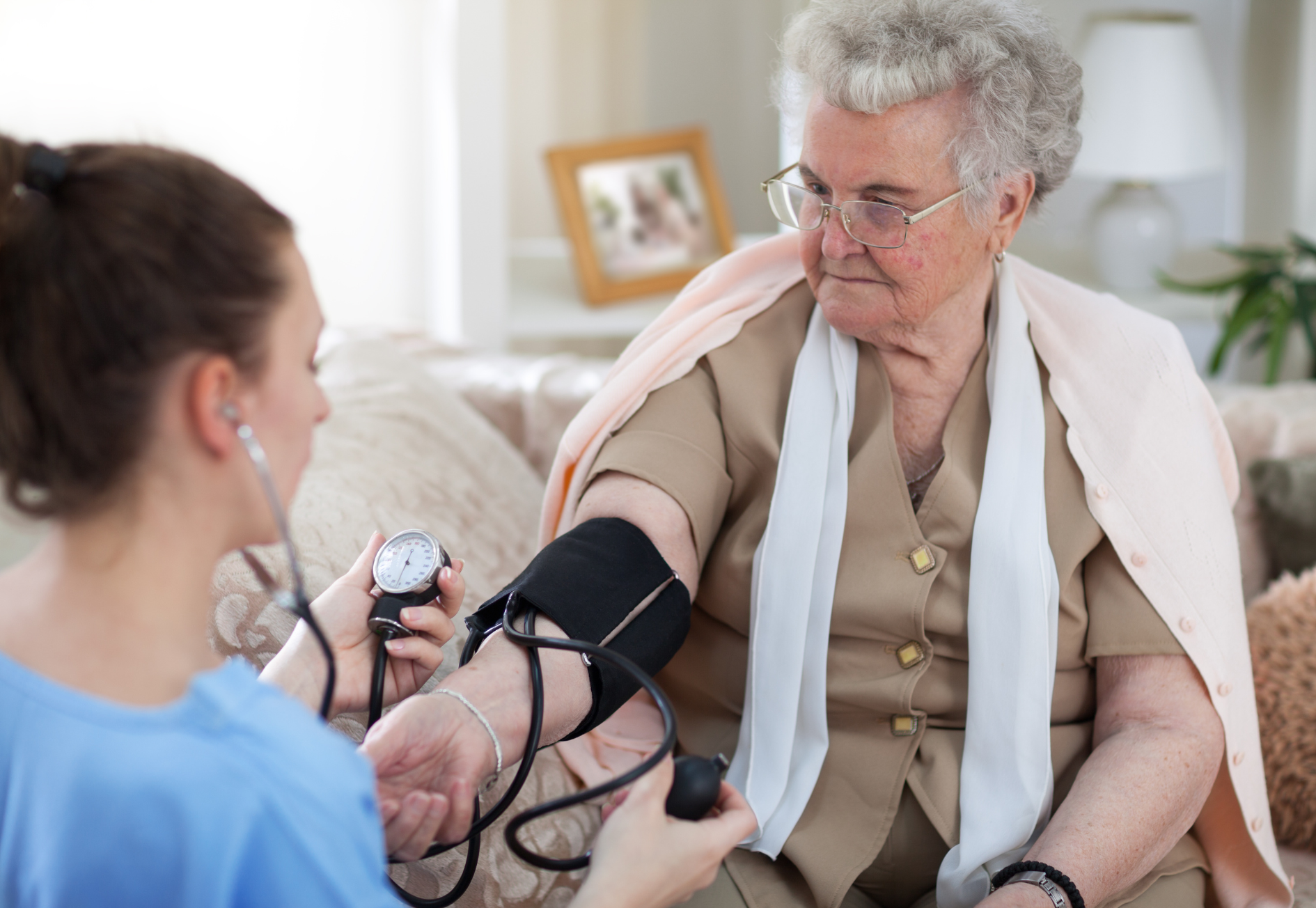 A healthcare worker checks an elderly woman's blood pressure in a living room.