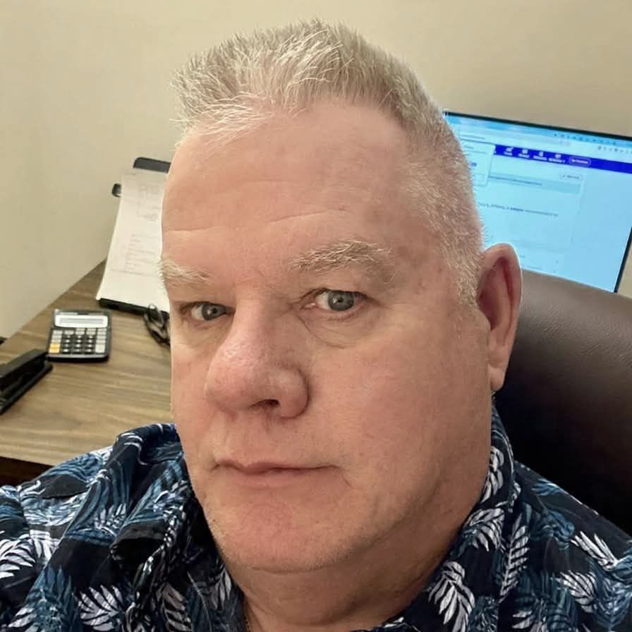 Man with gray hair, blue patterned shirt, looks at the camera in an office setting with a computer and desk supplies.