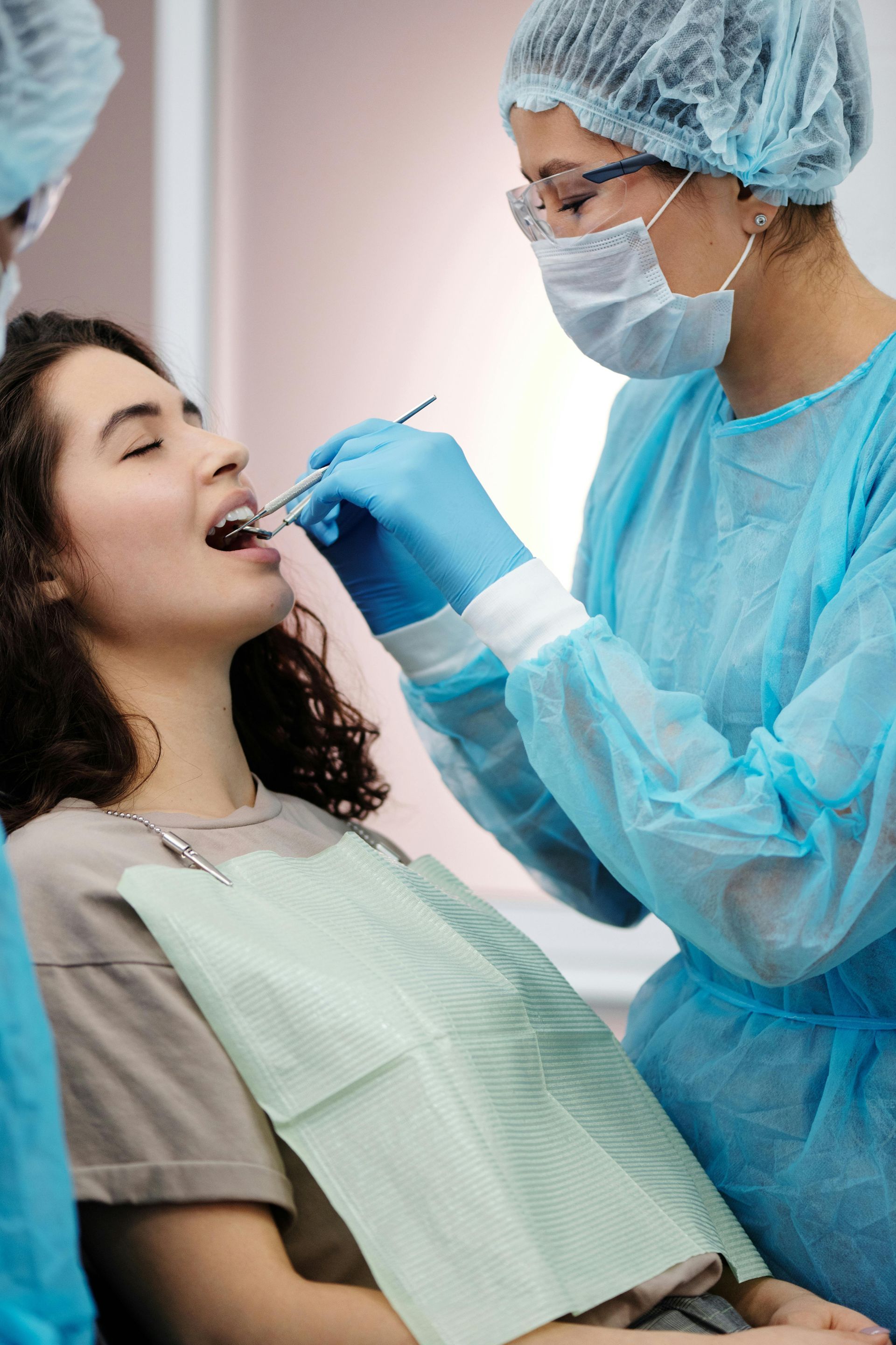 Dentist examining a patient's teeth. The patient reclines in a dental chair. Dentist wears a mask, gloves, and scrubs.