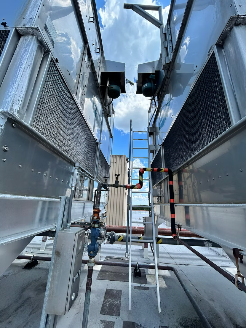 Cooling towers on a rooftop. Gray metal structures with a ladder and piping against a blue sky.
