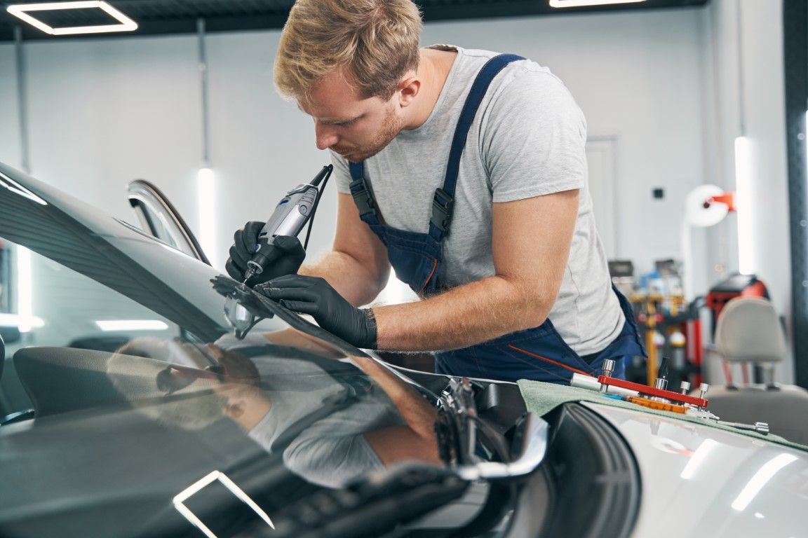 Mechanic in overalls repairs a car windshield with a handheld tool, indoors.