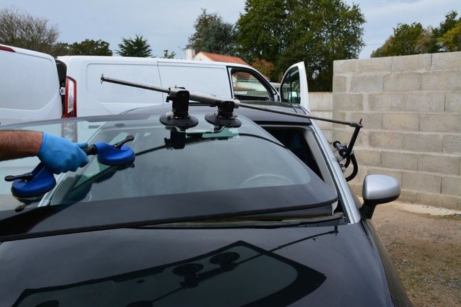 Person using suction cups to remove a car windshield in front of a van and brick wall.