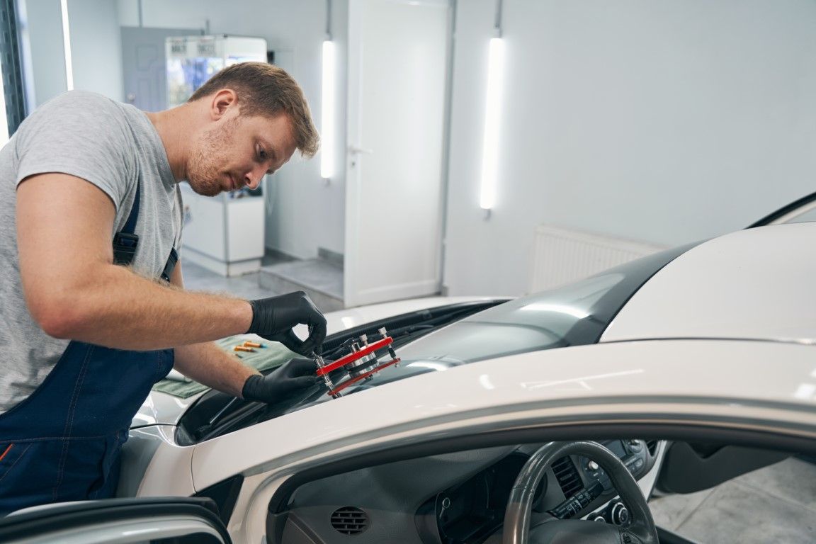 Mechanic repairing windshield on a white car, using tools in a garage.