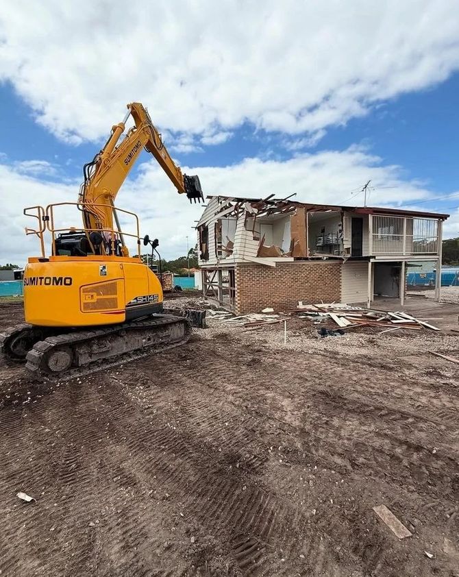A yellow excavator demolishing a two-story building on a brown dirt lot under a cloudy blue sky.