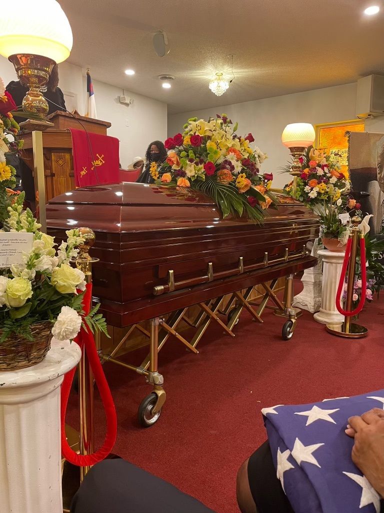A casket surrounded by flowers inside a church, with a person holding a folded American flag in the foreground.