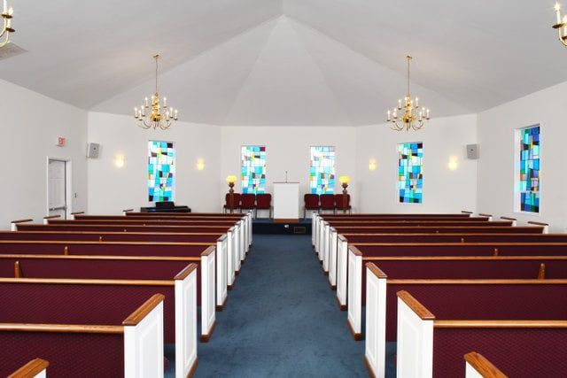 Interior of a chapel with rows of red pews, blue carpet, and stained glass windows.