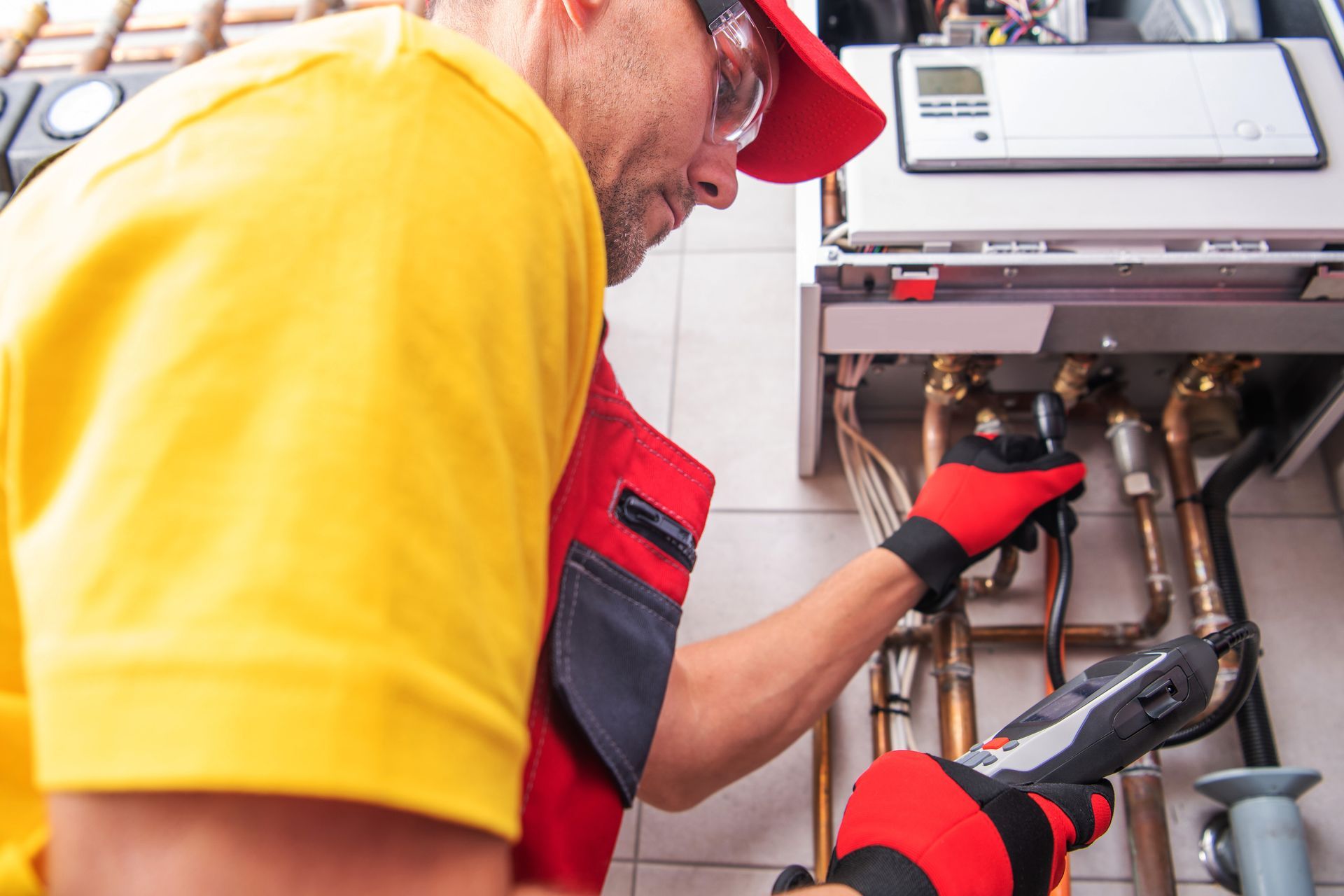 A man in a yellow shirt and red hat is working on a boiler.