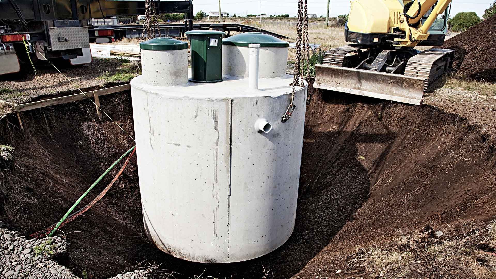 A concrete septic tank being lowered into a large hole by a crane near an excavator.