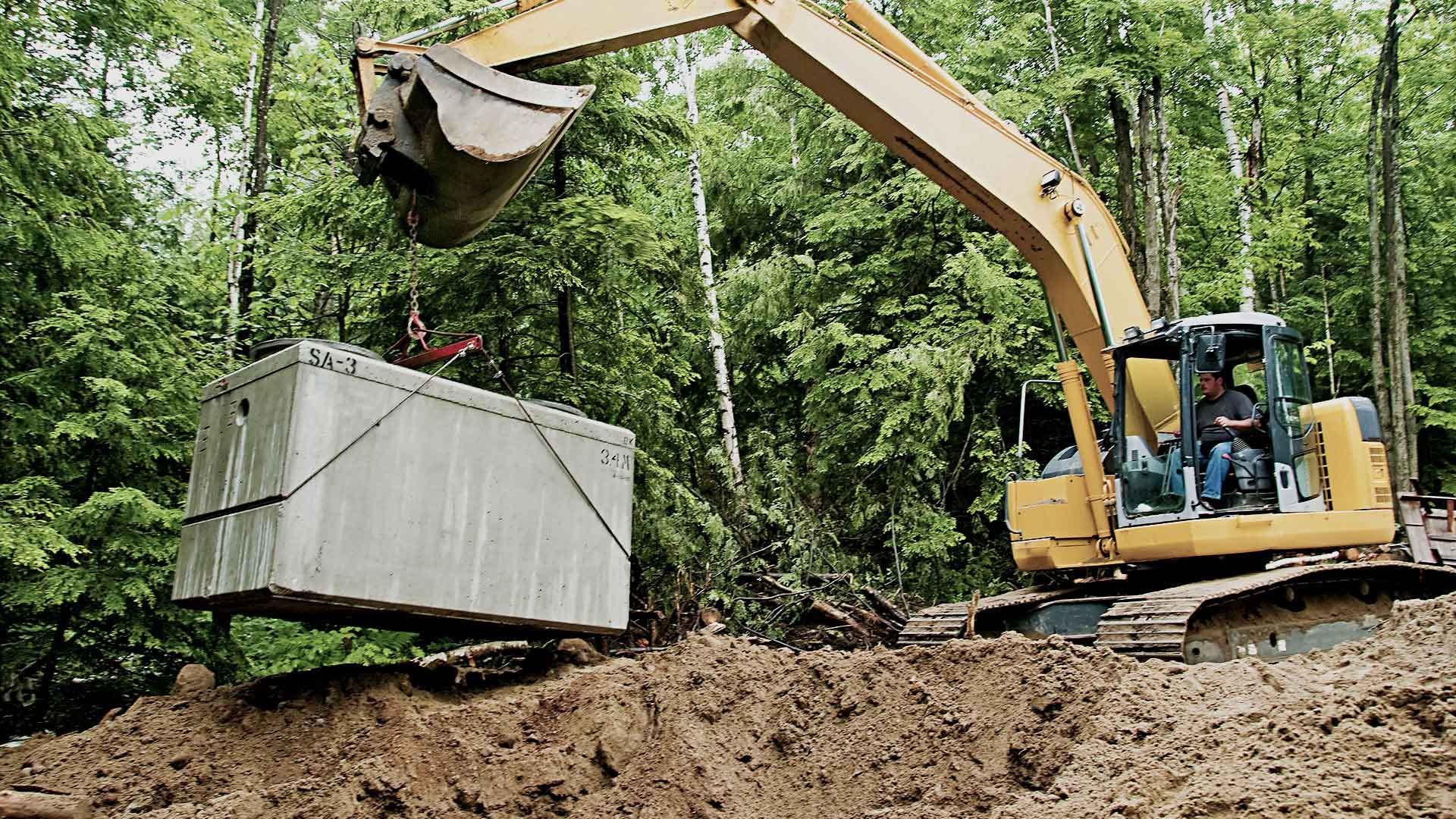 Yellow excavator lifting a large, rectangular concrete tank in a wooded area.