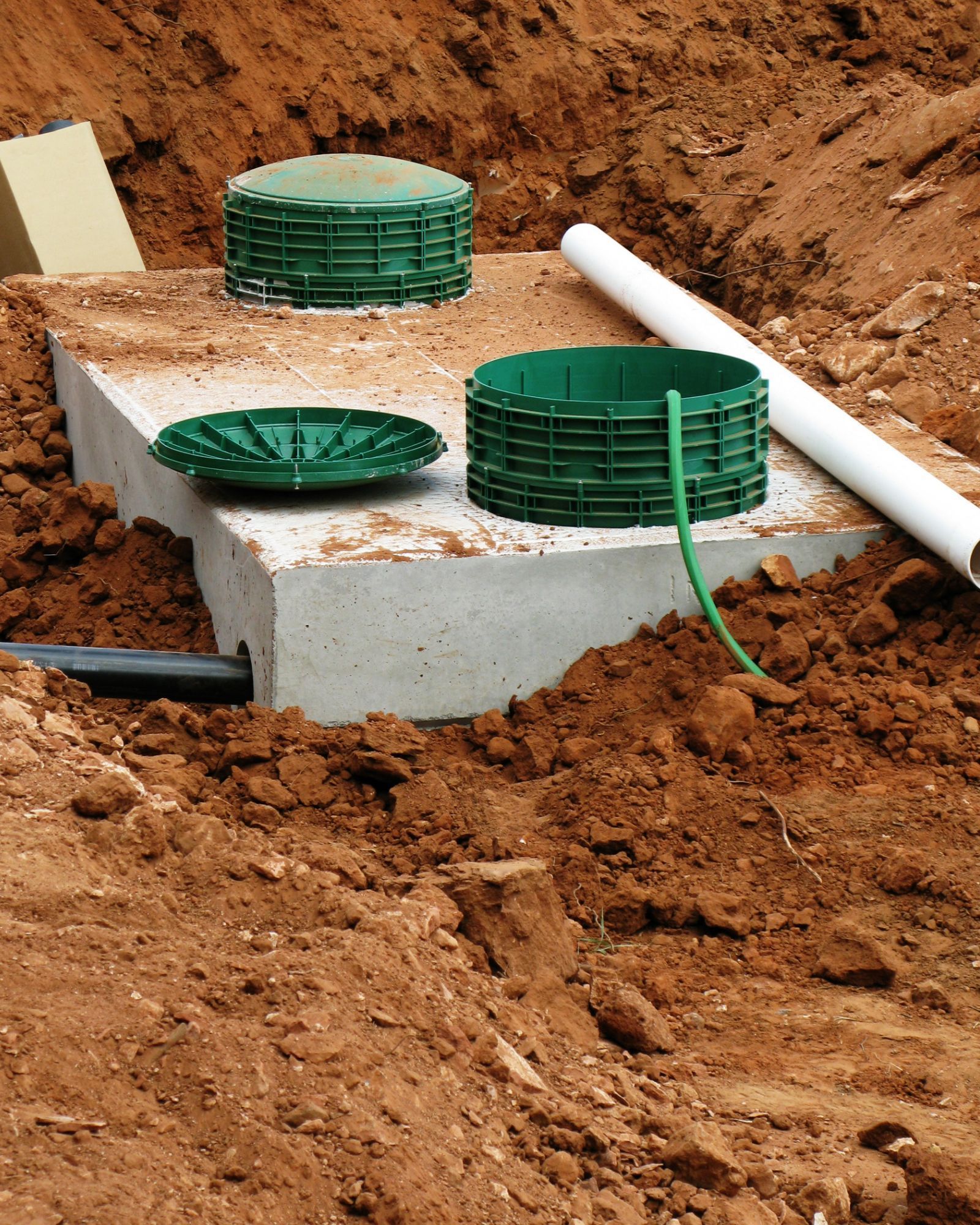 Concrete septic tank with green covers and pipes in a dirt excavation.
