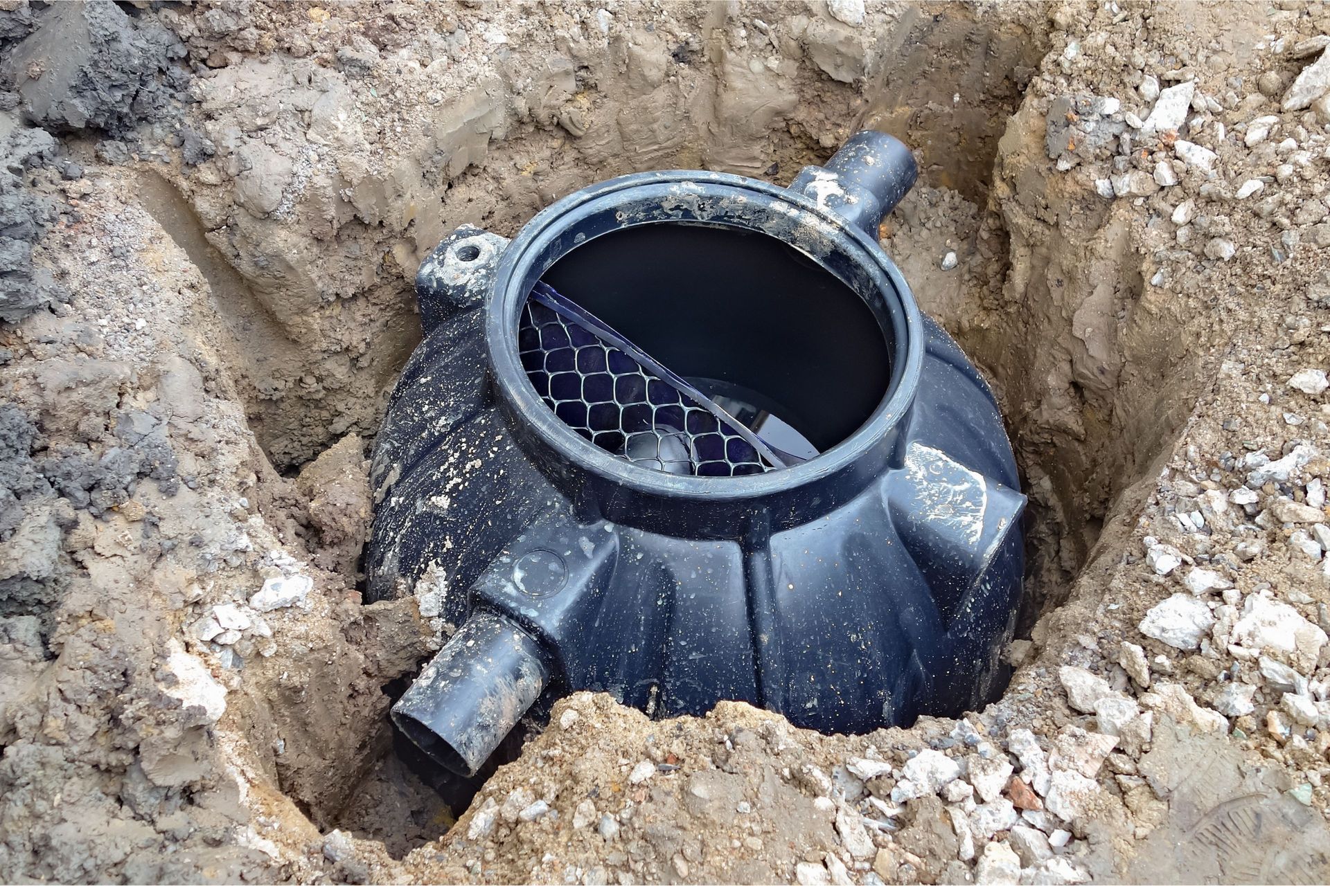 Black plastic water meter, partially buried in a trench, with open lid and connecting pipes.