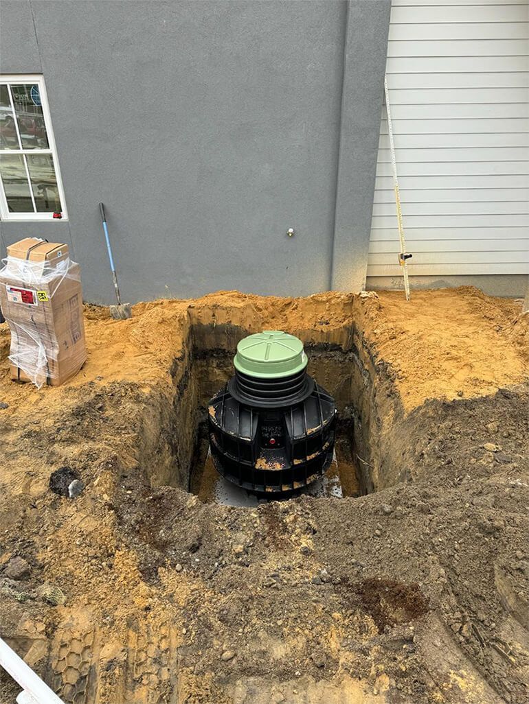 A black septic tank in a square hole in the ground, next to a building and partially covered with dirt.