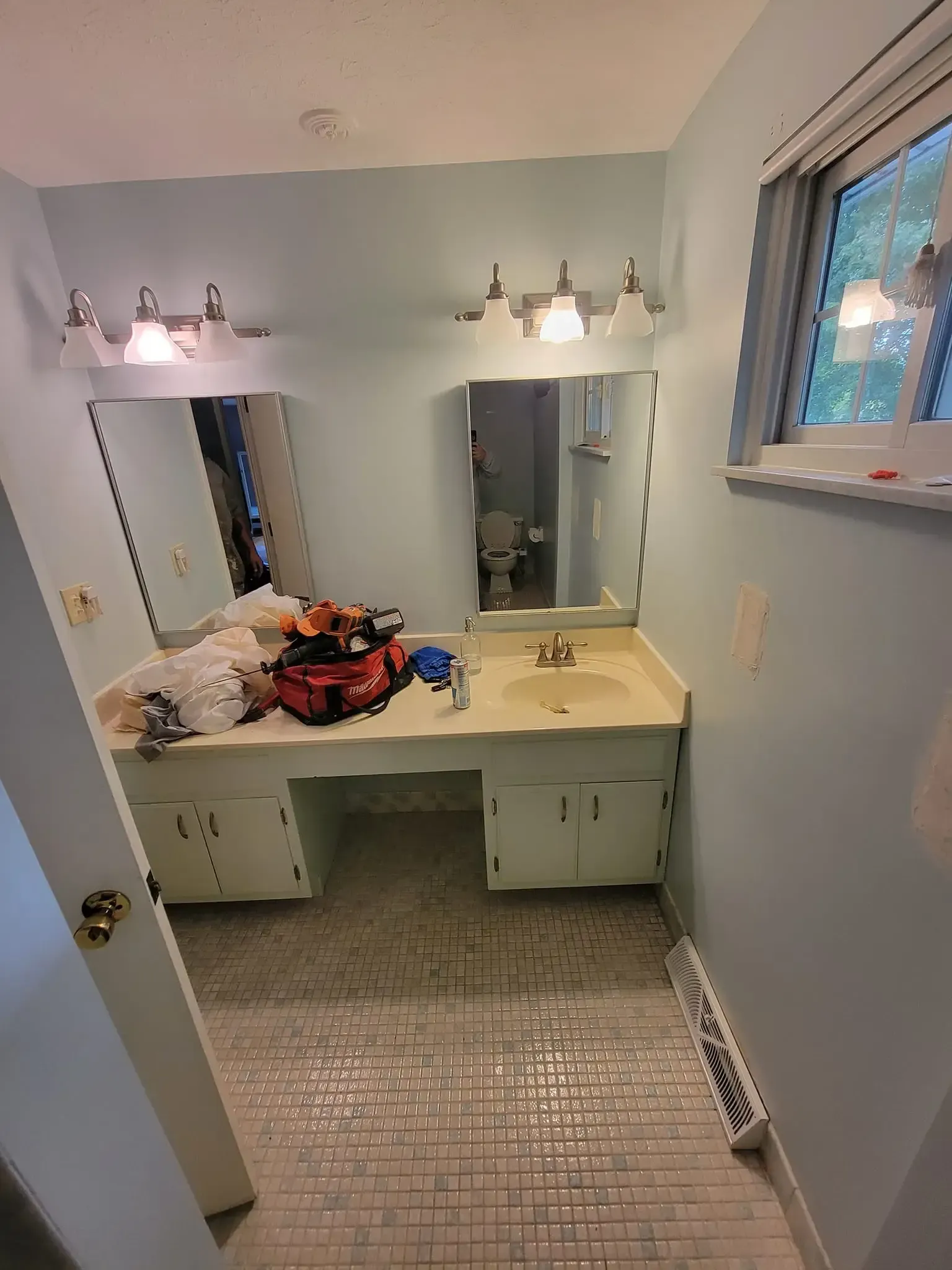 Bathroom with light blue walls, white vanity, two mirrors, and a window; tools are on the counter.