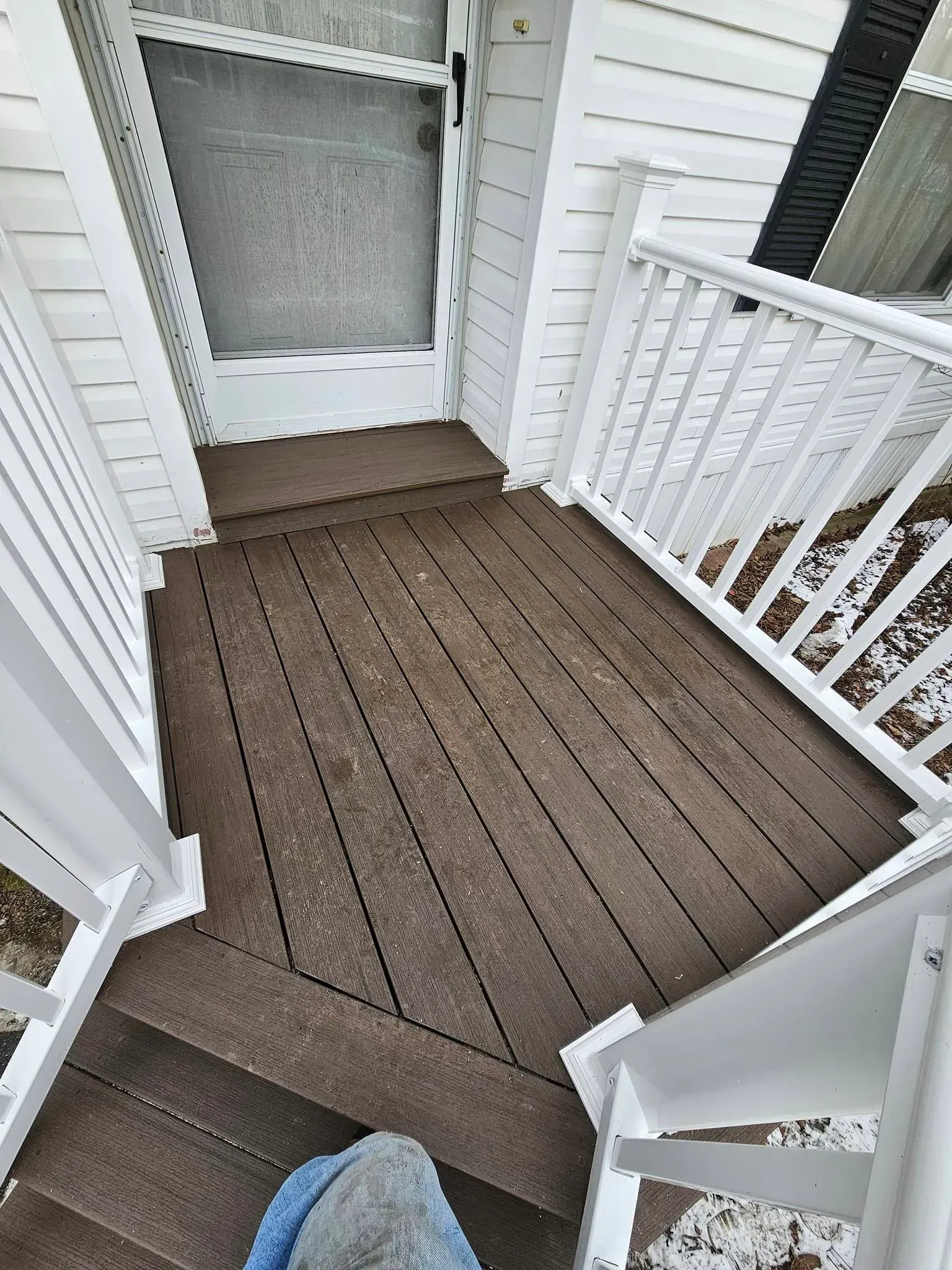 View of a small deck with stairs, brown decking, white railing and trim; snow on the ground.