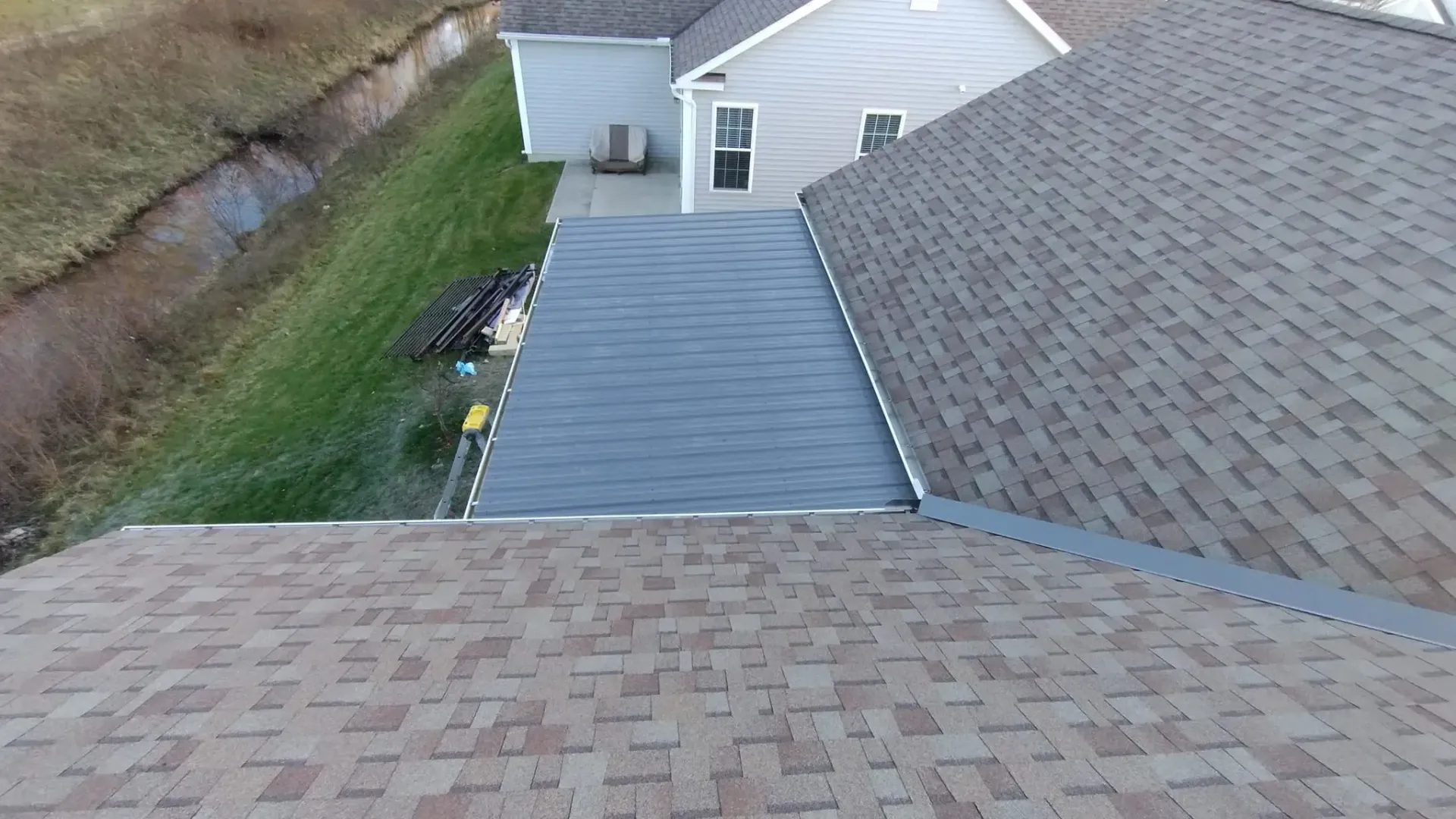 Overhead view of two brown shingled roofs, a gray flat roof, and a grassy yard.