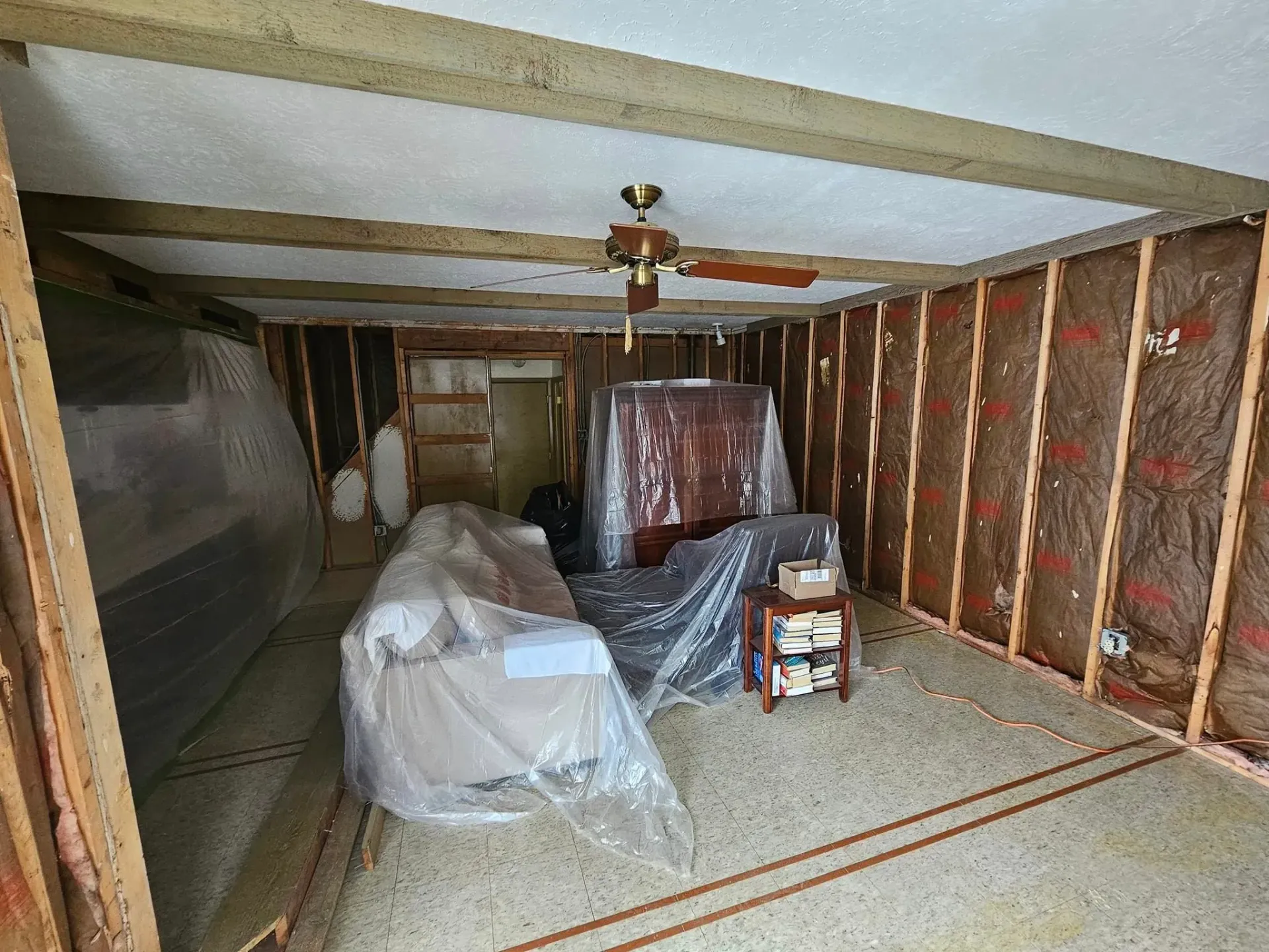 Room interior under renovation, exposed studs, insulation, furniture covered in plastic.
