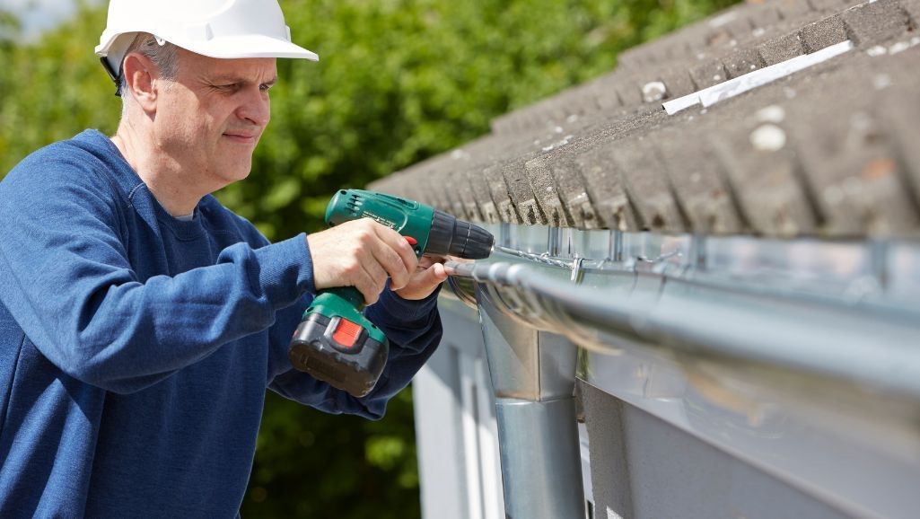 A man is installing a gutter on a roof with a drill.