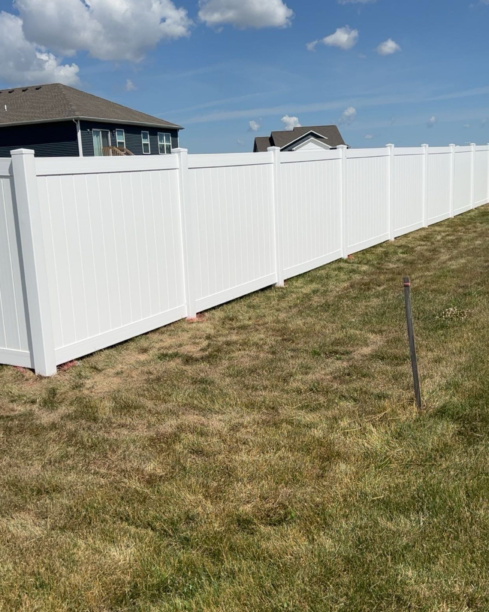 White vinyl fence on a slight incline, with houses and blue sky visible in the background.