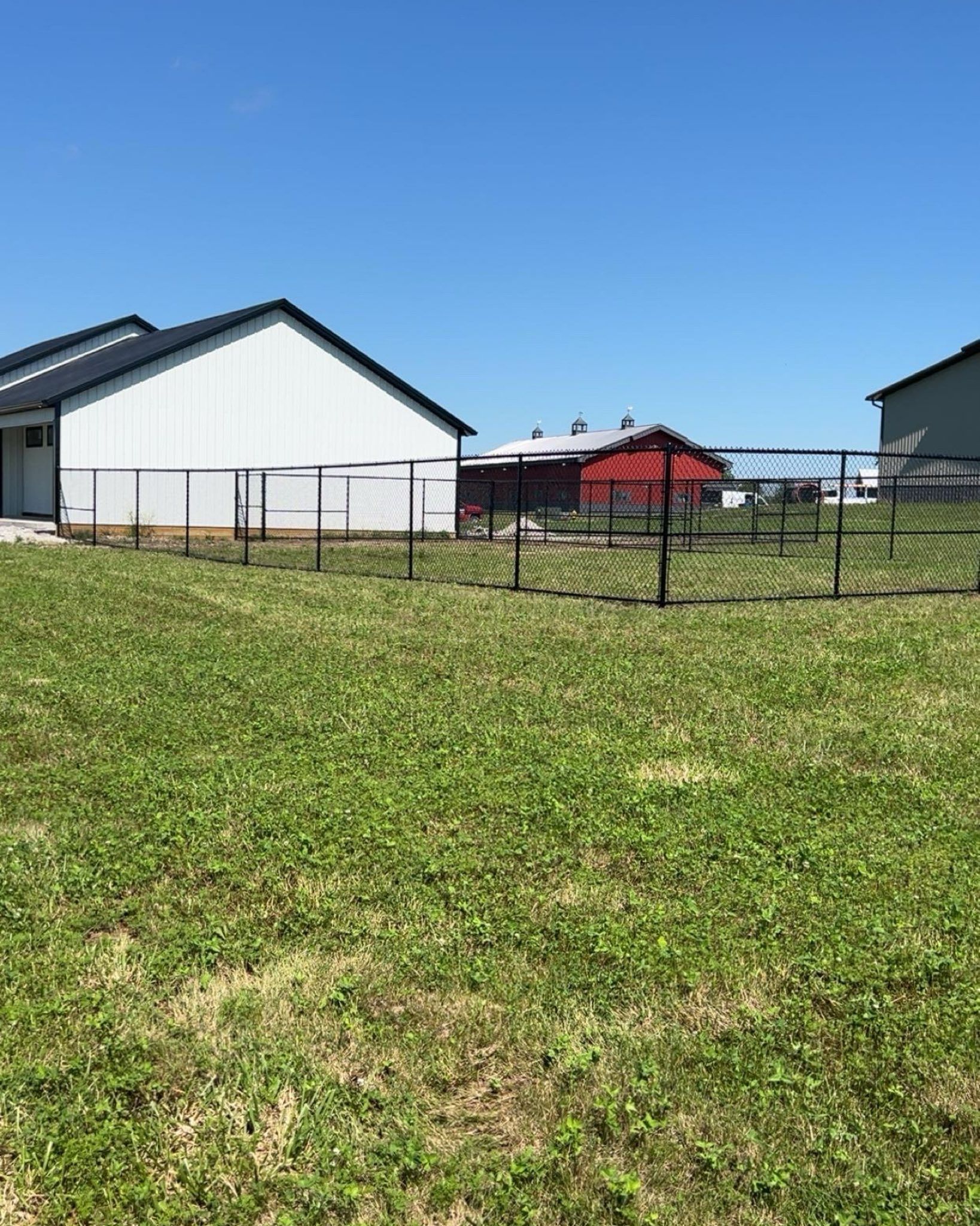 A green field with a black fence surrounding a white and red barn under a blue sky.