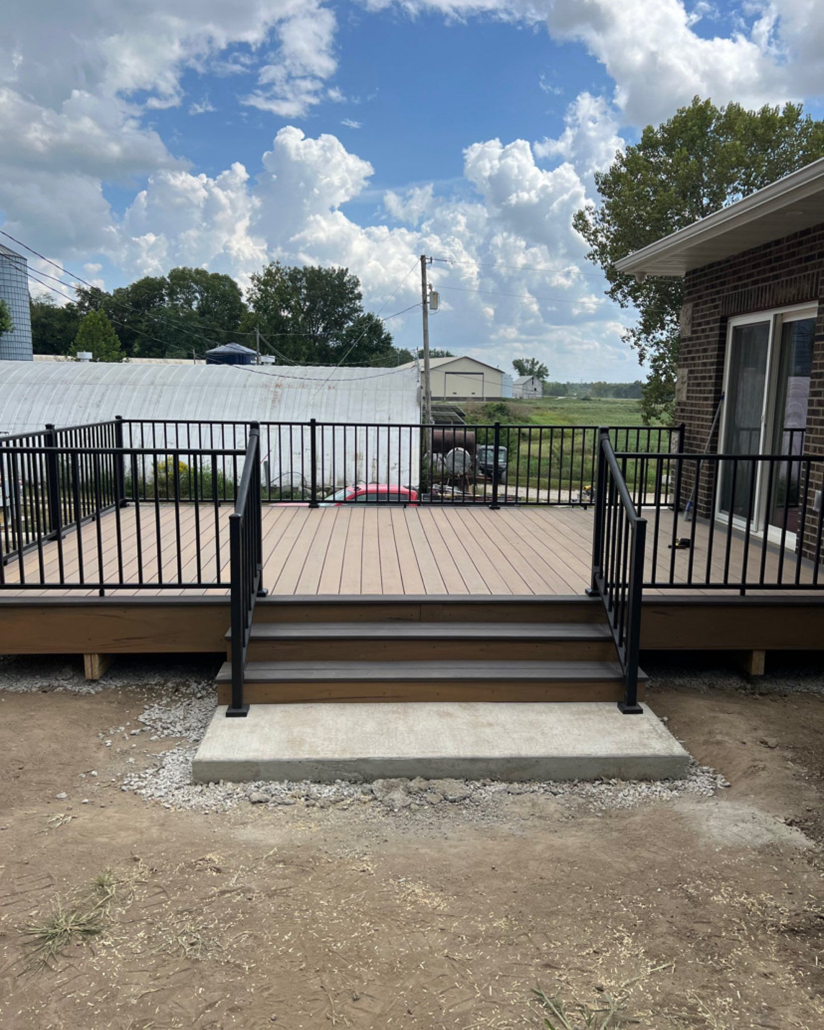 Wooden deck with black railings and steps, gray foundation, and a house, under a cloudy blue sky.