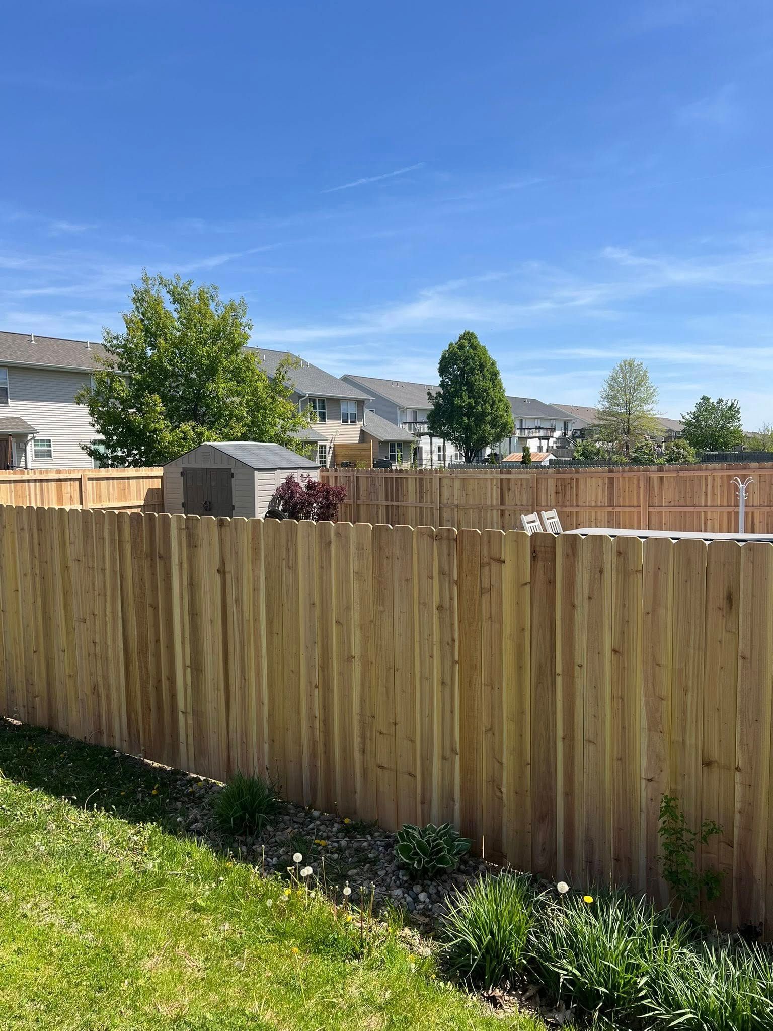 Wooden fence encloses a backyard with grass, shrubs, and a shed, under a blue sky.